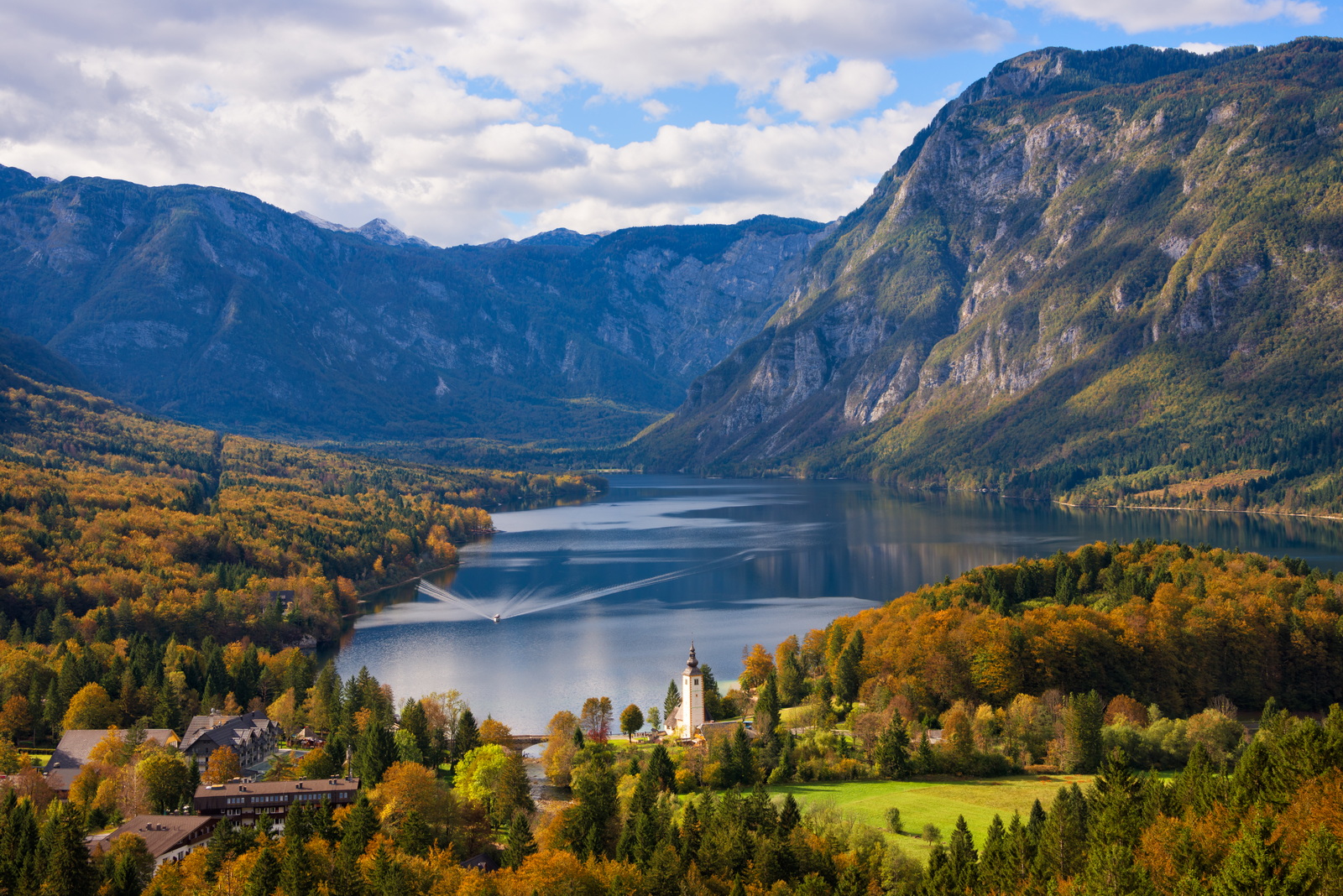 A ferry boat approaching the town of Ribčev Laz with the prominent church at the lakeside of Lake Bohinj surrounded by trees with foliage in autumn colors and the big Julian Alps mountains in Triglav National Park in a great fall landscape near Bled in Slovenia.